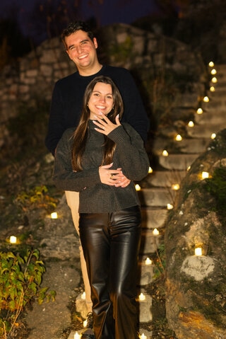 A happy couple celebrating their engagement proposal on the Gellért Hill in Budapest, standing on a romantically lit stone staircase surrounded by candles {{brizy_dc_image_alt imageSrc=