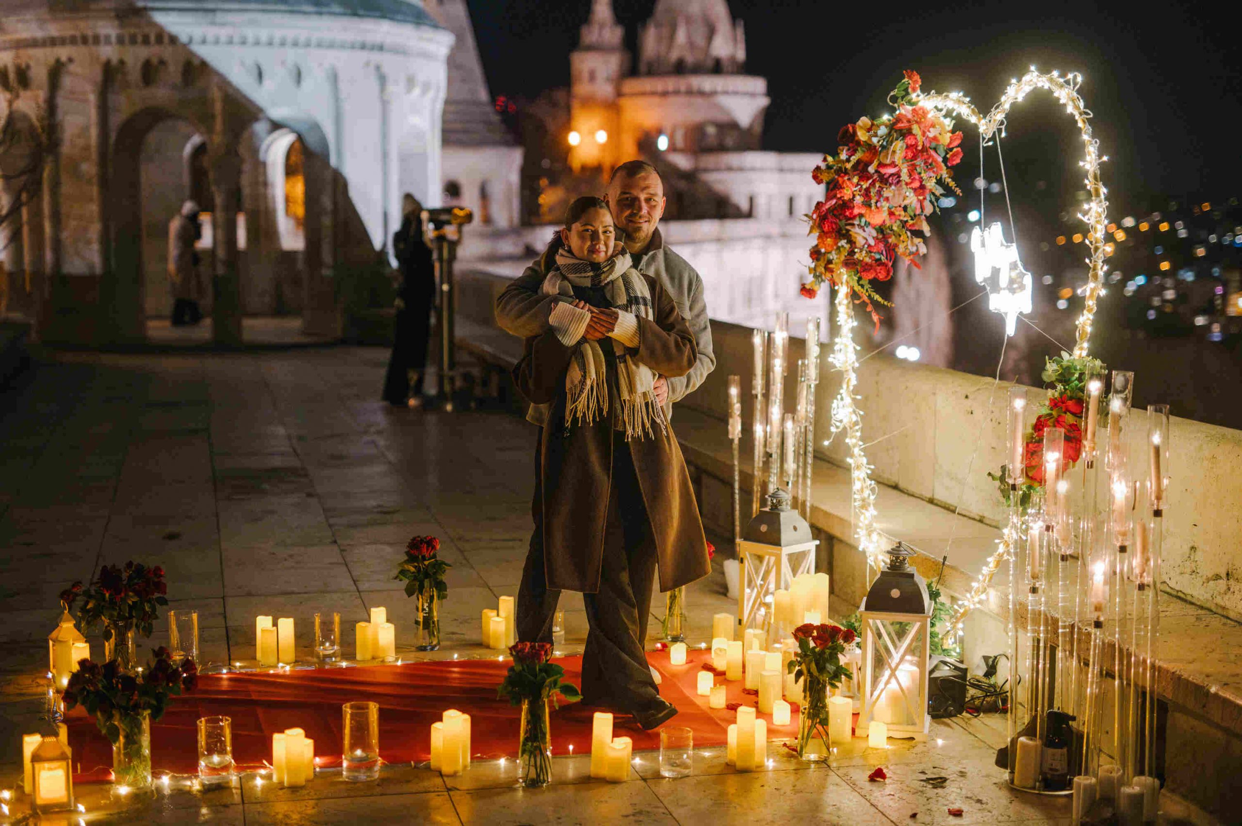 Couple embracing during a romantic proposal setup at Fisherman’s Bastion in Budapest, surrounded by candles, roses, and a glowing heart-shaped decoration {{brizy_dc_image_alt imageSrc=