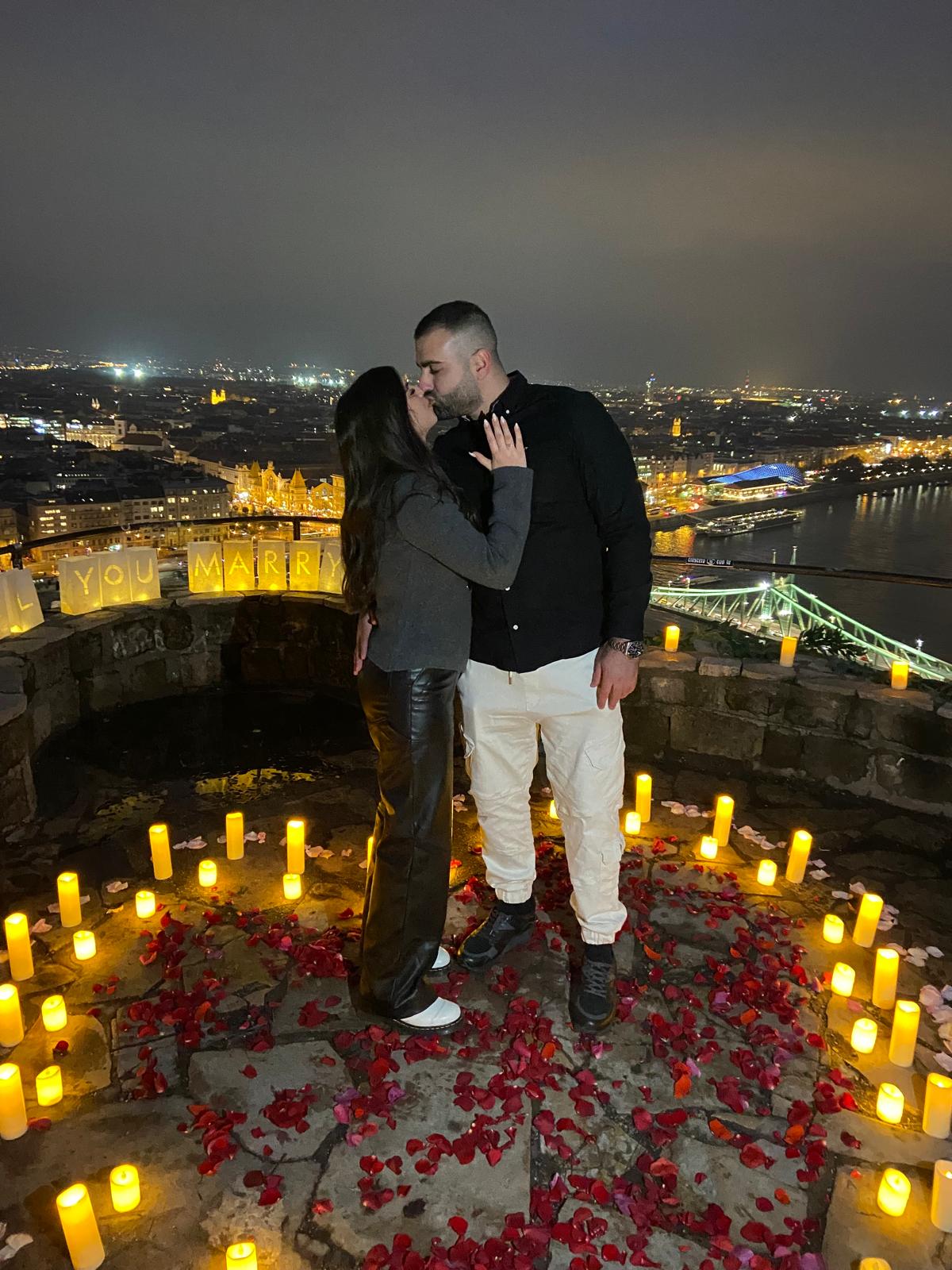 Romantic proposal on Gellért Hill in Budapest, surrounded by candles and rose petals, overlooking the city lights at night {{brizy_dc_image_alt imageSrc=