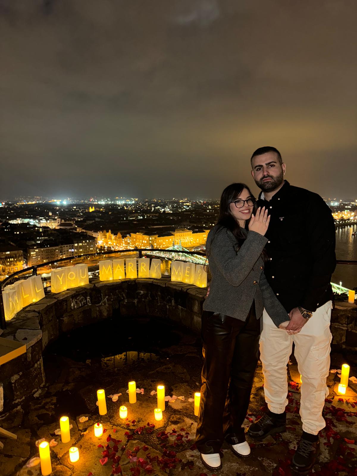 Romantic proposal on Gellért Hill in Budapest, surrounded by candles and rose petals, overlooking the city lights at night {{brizy_dc_image_alt imageSrc=