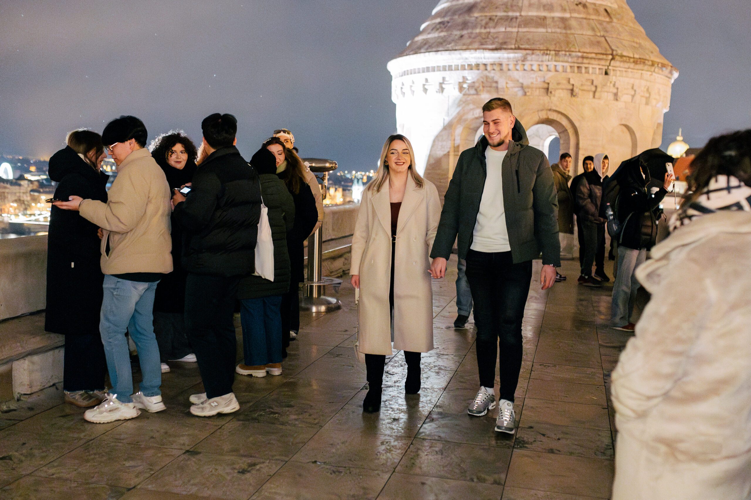 A Bosnian couple walking hand in hand at Fisherman’s Bastion in Budapest at night, surrounded by a crowd. The historic architecture and city lights create a magical atmosphere {{brizy_dc_image_alt imageSrc=