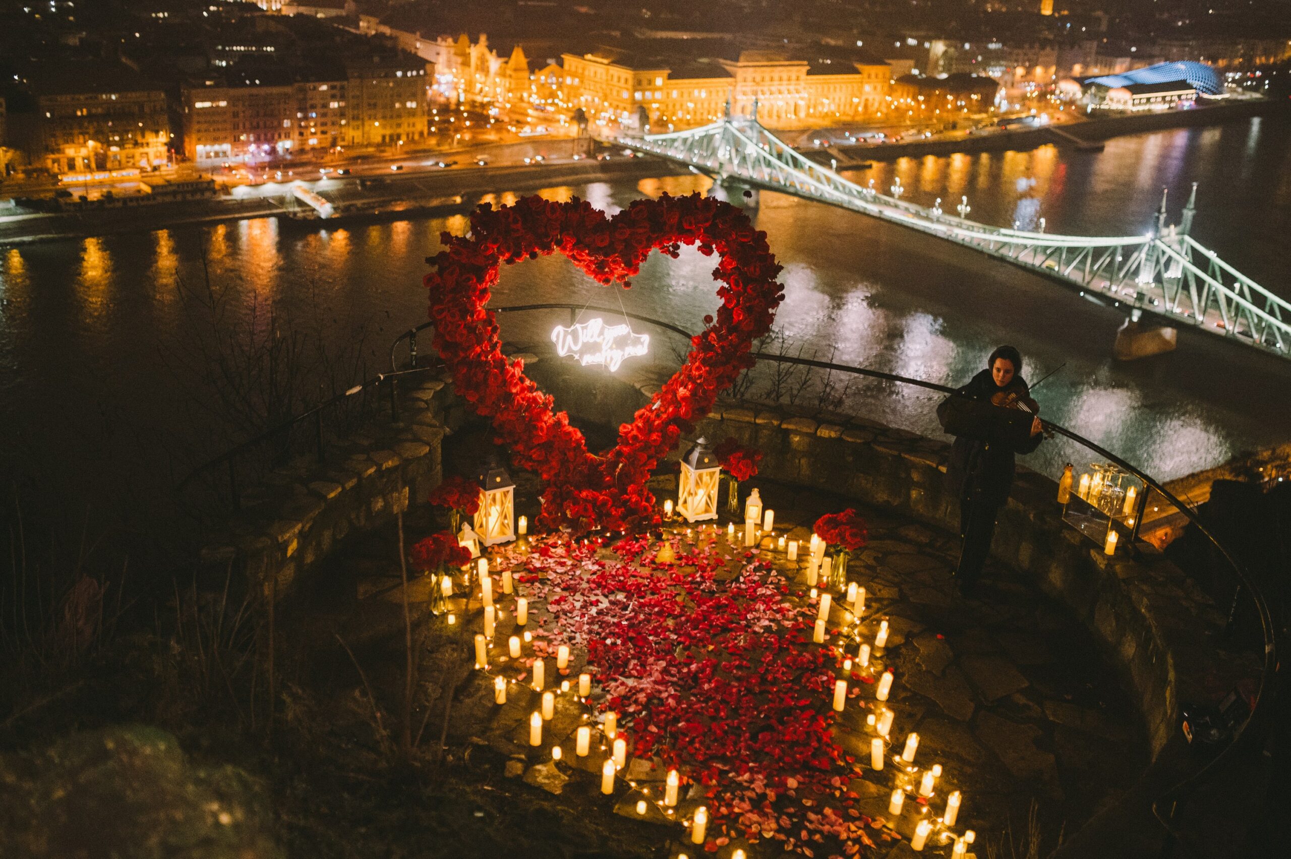 A breathtaking wedding proposal setup in Hungary, featuring a heart-shaped red rose arch, glowing candles, and a romantic cityscape in the background. {{brizy_dc_image_alt imageSrc=