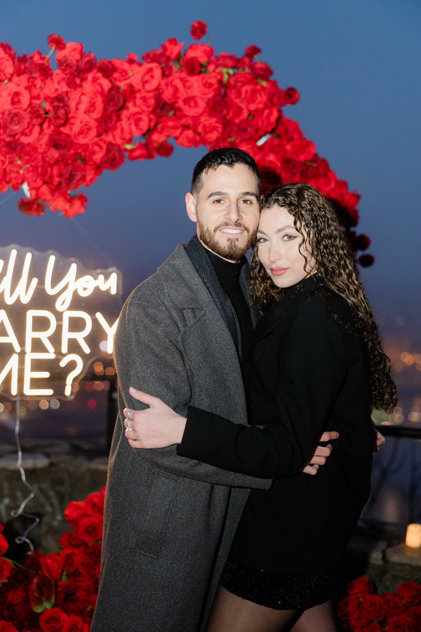 A Lebanese couple embracing in front of a romantic proposal setup with red roses and a neon sign that reads {{brizy_dc_image_alt imageSrc=