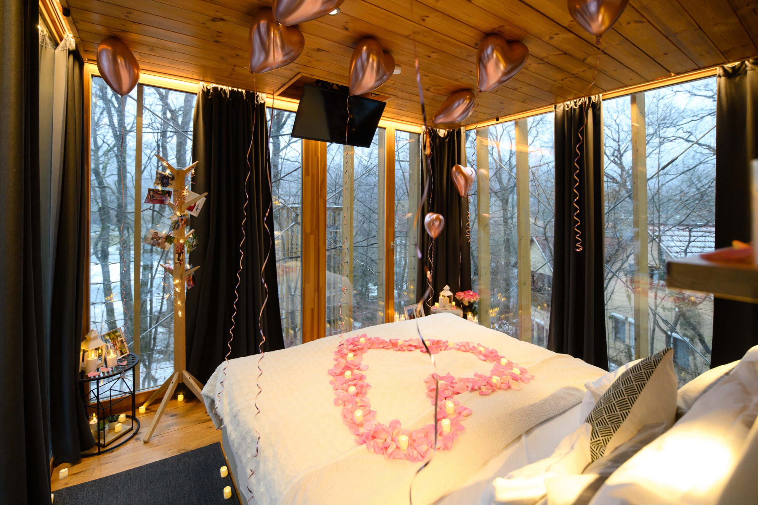 Cozy treehouse interior decorated for proposal with rose petal heart on white bed, rose gold balloons, fairy lights, and winter forest view through panoramic windows {{brizy_dc_image_alt imageSrc=