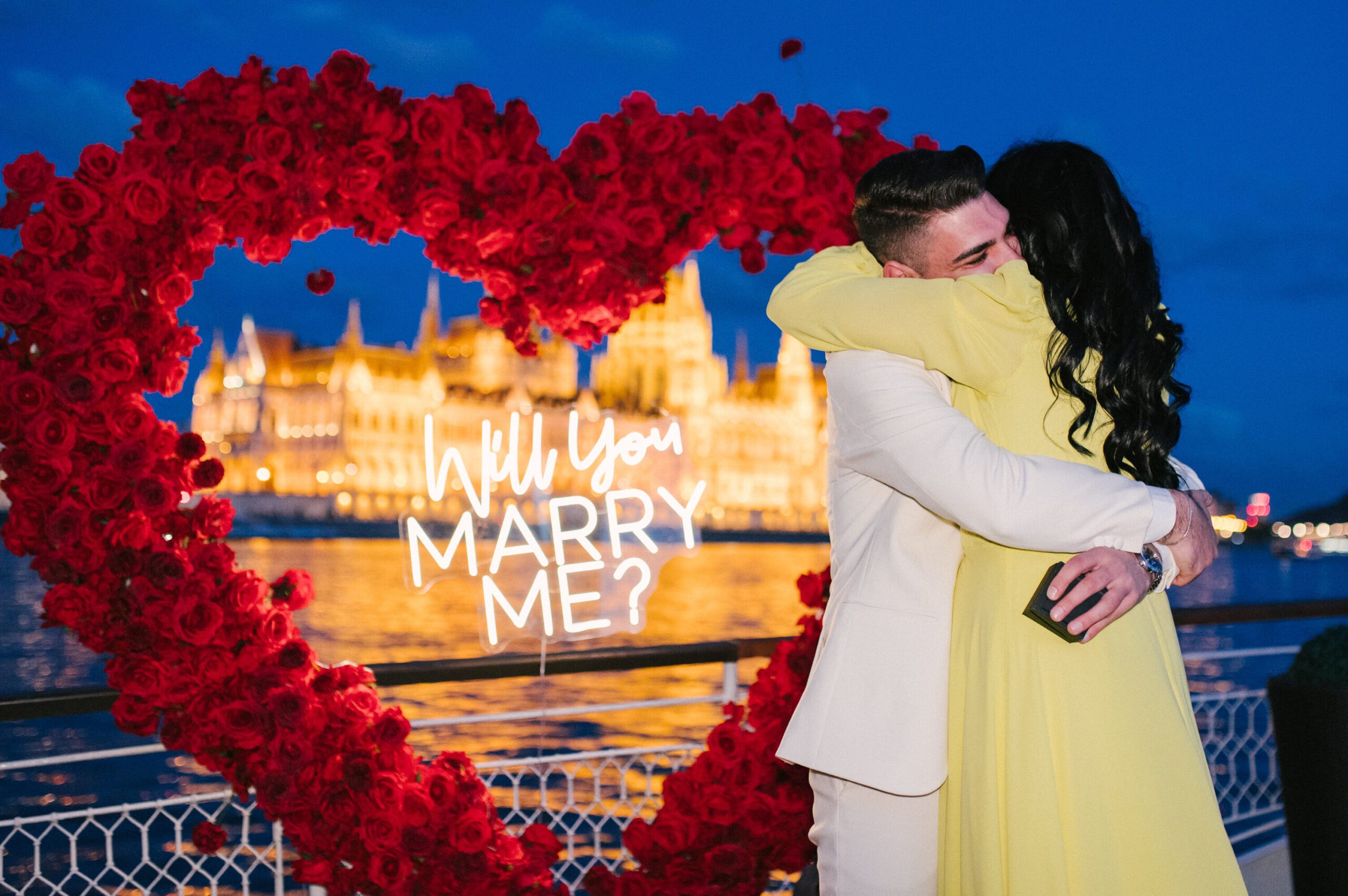Couple hugging during a romantic private boat proposal on the Danube in Budapest, with a heart-shaped rose arch and the illuminated Parliament in the background. {{brizy_dc_image_alt imageSrc=