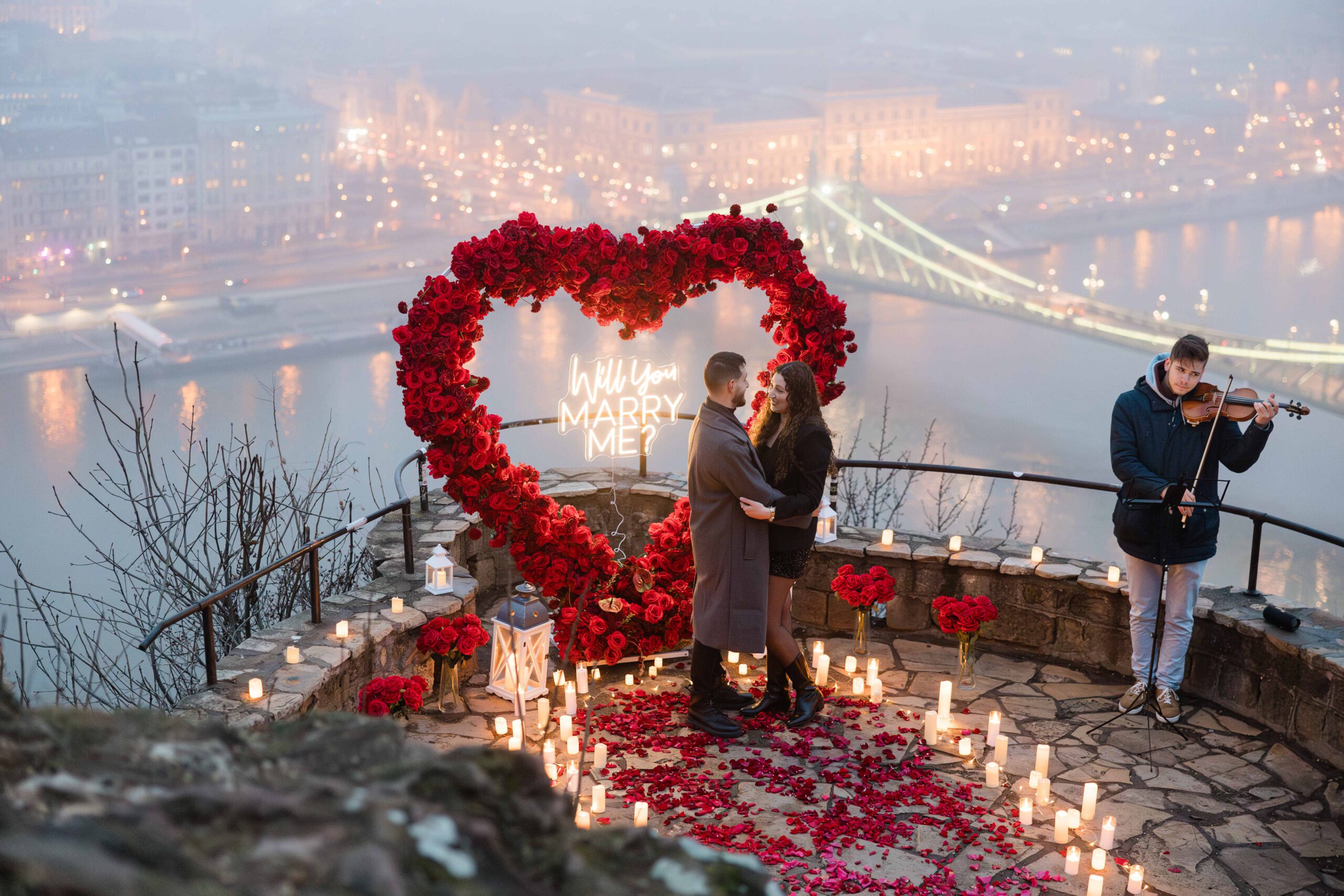 Engagement proposal with heart-shaped flower arch, candles, and live violin music overlooking Budapest {{brizy_dc_image_alt imageSrc=