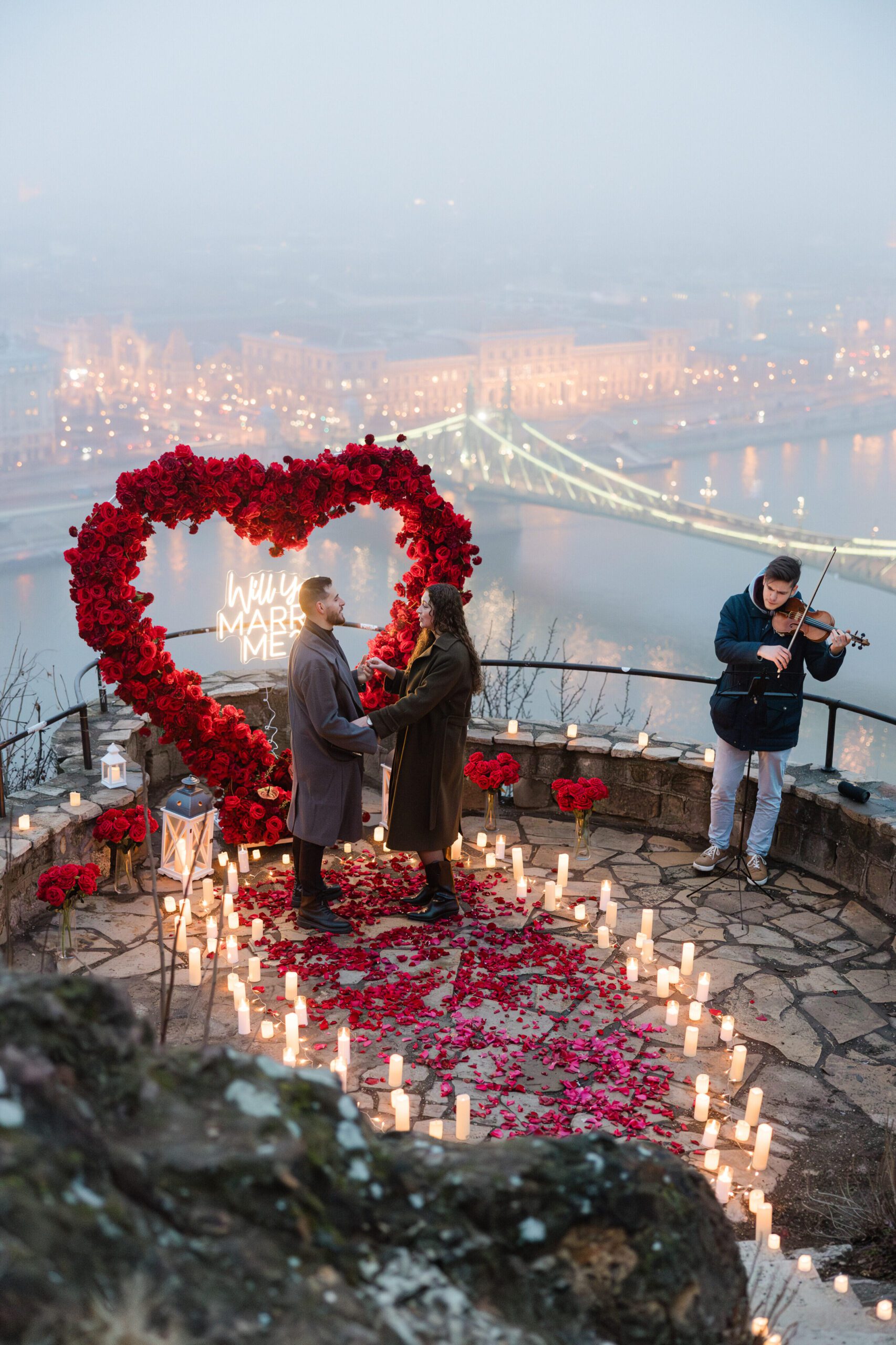 Romantic marriage proposal with red rose heart, candlelight, and violinist on panoramic viewpoint in Budapest {{brizy_dc_image_alt imageSrc=