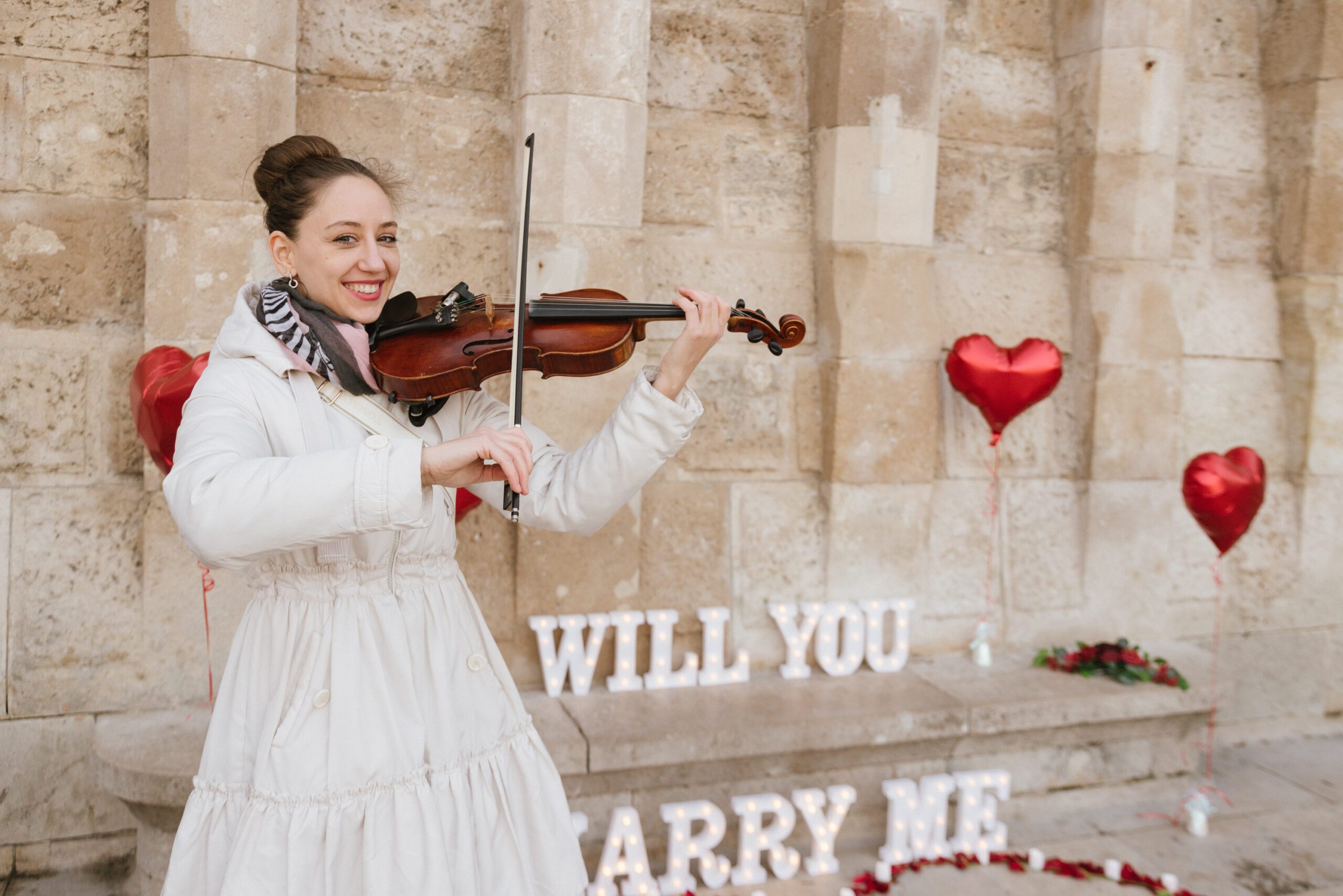 Female violinist playing romantic music during a surprise marriage proposal in Budapest with red heart balloons and glowing {{brizy_dc_image_alt imageSrc=