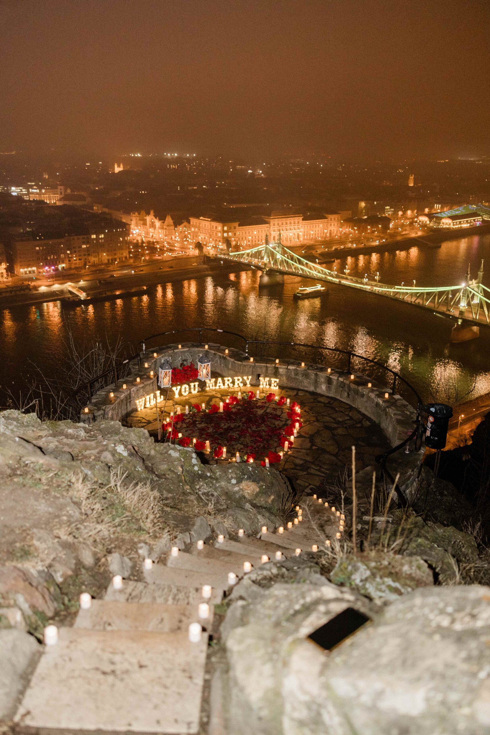 Wedding proposal setup with candles and rose petals overlooking Budapest and Liberty Bridge at night {{brizy_dc_image_alt imageSrc=