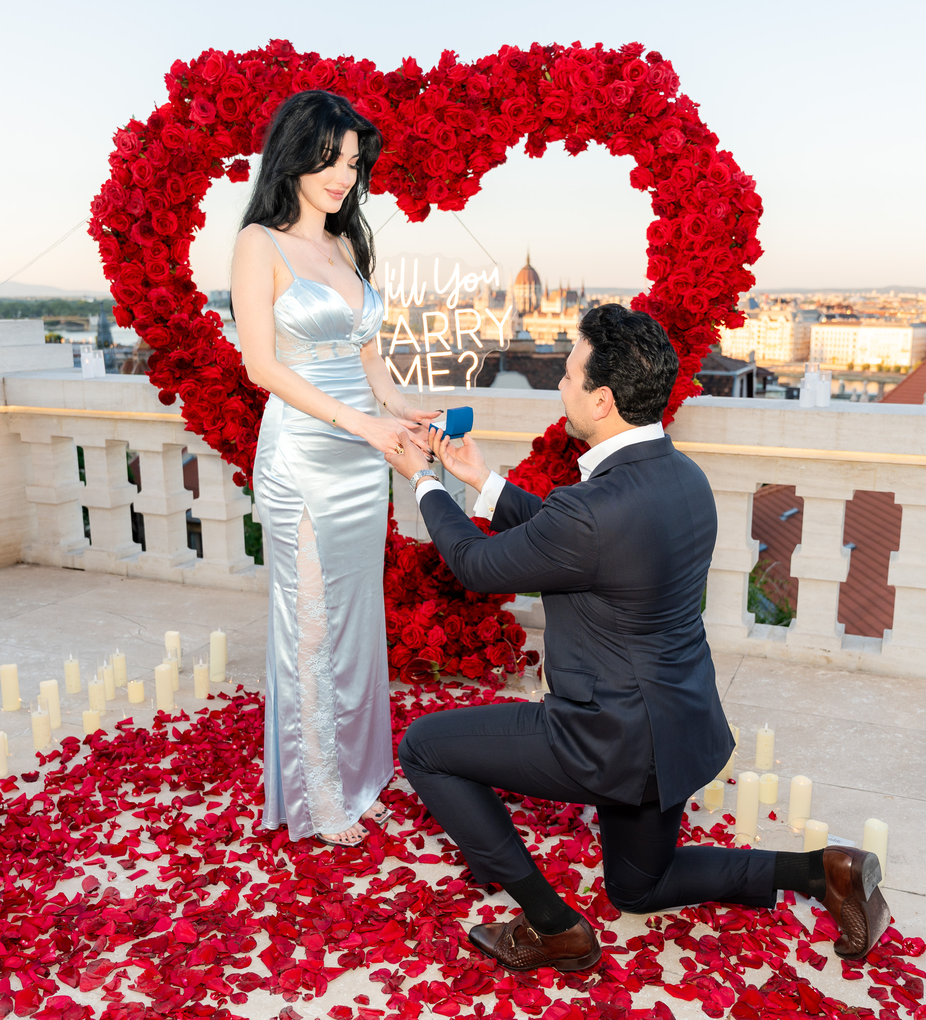 Elegant engagement on a Budapest terrace with rose petals, candles, and heart-shaped floral arch {{brizy_dc_image_alt imageSrc=