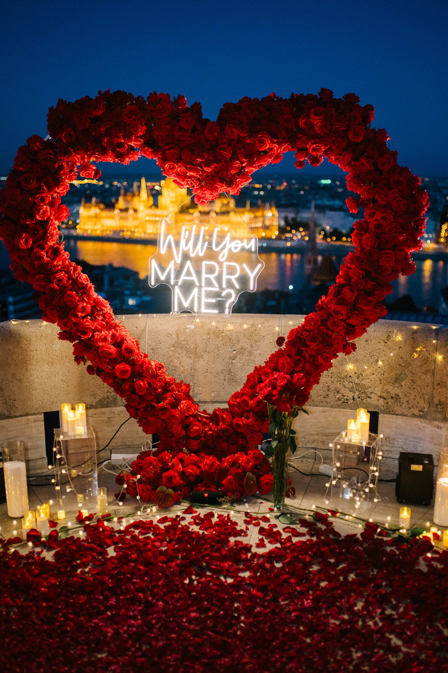 Luxury marriage proposal setup with a red rose heart, candles and “Will You Marry Me?” sign overlooking the Liberty Bridge in Budapest {{brizy_dc_image_alt imageSrc=