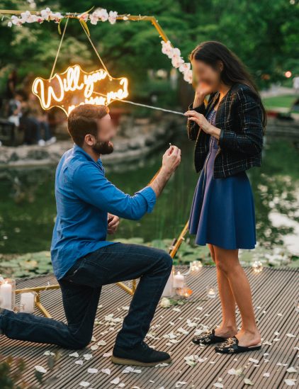 A man proposing to a woman at Margaret Island with a neon sign reading 'Will you marry me?' in the background.