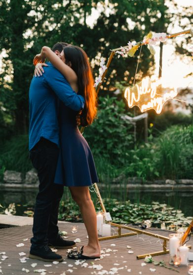 A couple hugging after a proposal with a 'Will you marry me?' neon sign at the Japanese Garden in Margaret Island, Budapest, with a pond in the background.