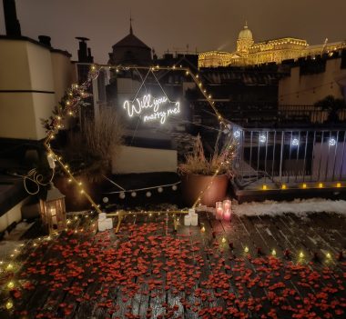 Romantic_Budapest_Rooftop_Proposal with Candlelit Table, Scattered Rose Petals, Champagne, and 'Will_You_Marry_Me' Neon Sign against City Skyline