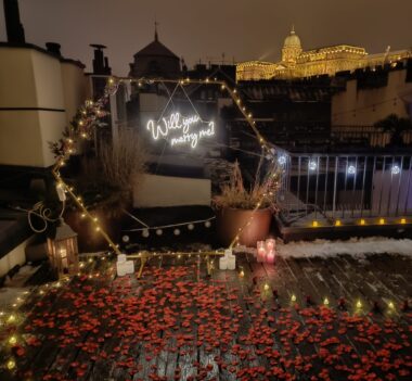 Romantic_Budapest_Rooftop_Proposal with Candlelit Table, Scattered Rose Petals, Champagne, and 'Will_You_Marry_Me' Neon Sign against City Skyline {{brizy_dc_image_alt imageSrc=
