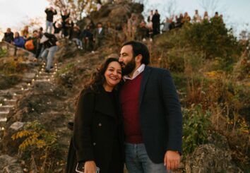 A joyful couple shares a loving moment after an engagement, surrounded by nature and a gathering of well-wishers on a hillside {{brizy_dc_image_alt imageSrc=