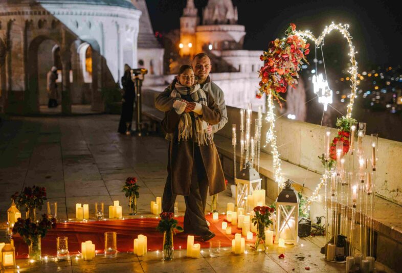 Couple embracing during a romantic proposal setup at Fisherman’s Bastion in Budapest, surrounded by candles, roses, and a glowing heart-shaped decoration {{brizy_dc_image_alt imageSrc=