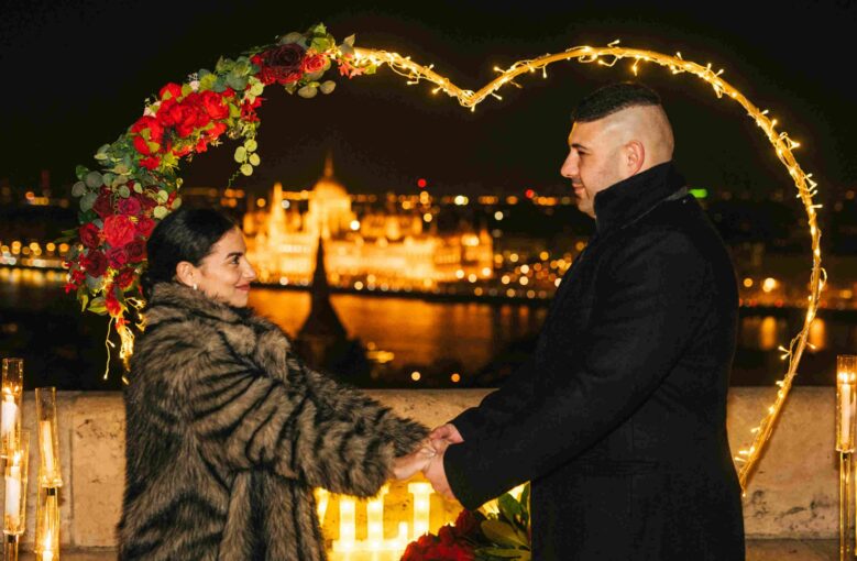 Couple holding hands under a heart-shaped floral decoration in Budapest with the Parliament building lit up at night {{brizy_dc_image_alt imageSrc=