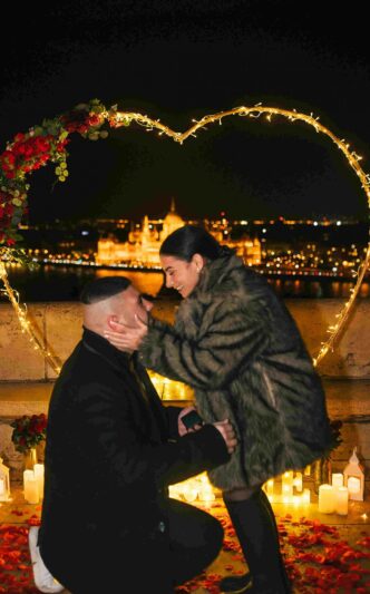 Man on one knee proposing to his partner under a heart-shaped light decoration in Budapest with the Parliament building glowing in the background {{brizy_dc_image_alt imageSrc=