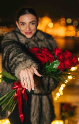 Smiling woman showing her engagement ring, holding red roses with Budapest’s romantic city lights in the background {{brizy_dc_image_alt imageSrc=