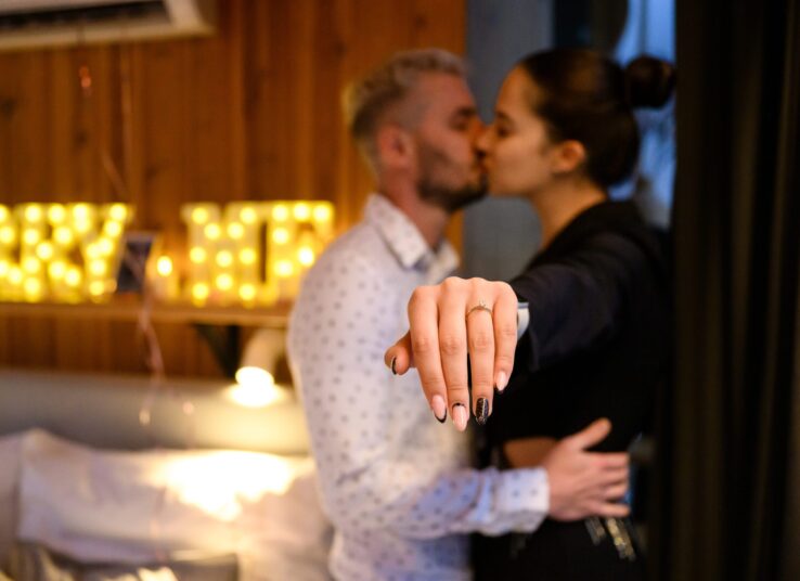 Első csók jegyespárként a Faház Hotel szobájában, fókuszban a gyémántgyűrűs kéz, háttérben fénydekoráció és romantikus hangulat {{brizy_dc_image_alt imageSrc=