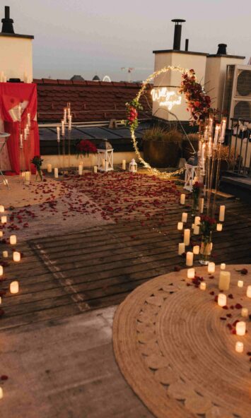 Romantic rooftop proposal setup in Budapest with rose petals, glowing candles, a floral arch with a 'Will you marry me?' neon sign, and elegant decorations {{brizy_dc_image_alt imageSrc=