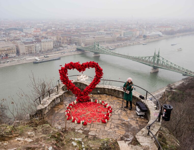Romantic proposal setup at Gellért Hill Budapest with large red rose heart arch and candles overlooking Liberty Bridge and Danube river, with violinist in green coat {{brizy_dc_image_alt imageSrc=