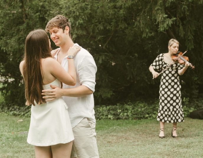 A couple embracing during a romantic marriage proposal while a violinist plays in the background on Margaret Island, Budapest.