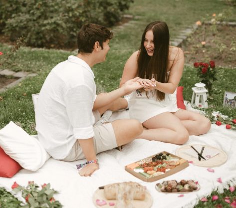 A couple sitting on a picnic blanket during a marriage proposal on Margaret Island, Budapest, as the man places a ring on the woman’s finger.