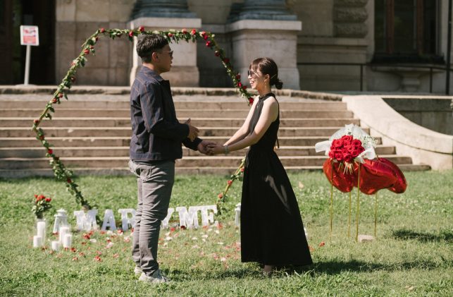 A couple holding hands during a marriage proposal in front of the historical Széchenyi Bath House in Budapest, with romantic decor including a heart-shaped rose arch.