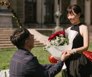 A man from Hong Kong on one knee, proposing with a bouquet of red roses to his partner in front of the Széchenyi Bath House in Budapest's city park