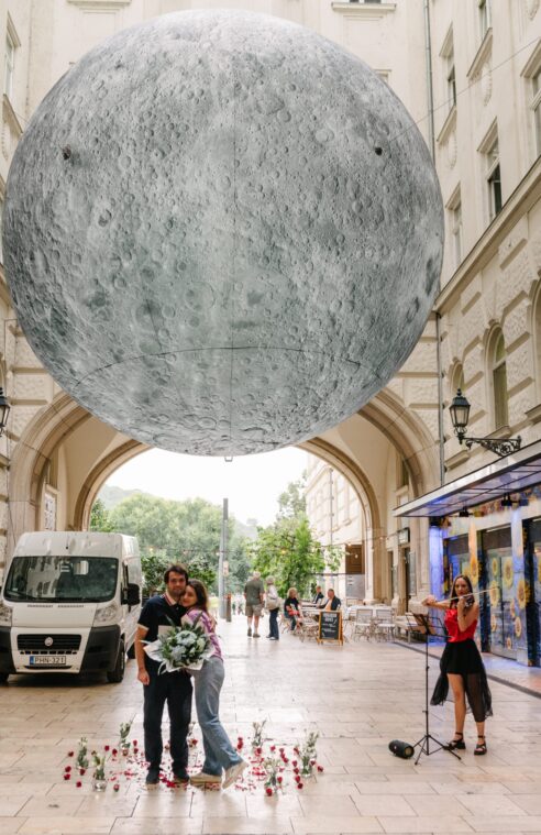 Couple celebrating their engagement under a large moon installation in Budapest's historic center, with a violinist playing nearby {{brizy_dc_image_alt imageSrc=