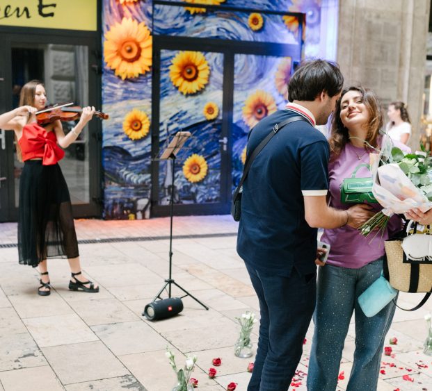 Couple sharing a romantic moment after a proposal in Budapest's historic center with a violinist playing in the background and a colorful sunflower mura