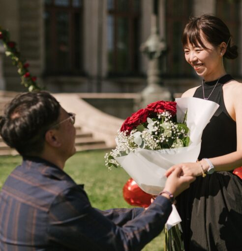 A man from Hong Kong on one knee, proposing with a bouquet of red roses to his partner in front of the Széchenyi Bath House in Budapest's city park {{brizy_dc_image_alt imageSrc=