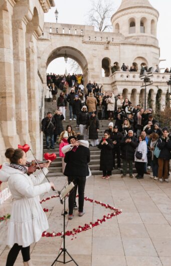 Public marriage proposal at Fisherman’s Bastion in Budapest with live violin music, red roses, and cheering crowd {{brizy_dc_image_alt imageSrc=