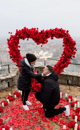 Will you marry me proposal setup at Budapest viewpoint with red rose heart arch, candles, and glowing neon sign {{brizy_dc_image_alt imageSrc=