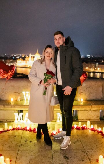 A newly engaged Bosnian couple poses in a romantic candlelit proposal setup at Fisherman’s Bastion in Budapest. The woman holds red roses, surrounded by glowing candles, rose petals, and a breathtaking city view. {{brizy_dc_image_alt imageSrc=