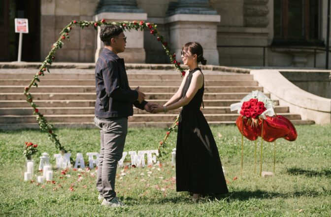 A couple holding hands during a marriage proposal in front of the historical Széchenyi Bath House in Budapest, with romantic decor including a heart-shaped rose arch. {{brizy_dc_image_alt imageSrc=