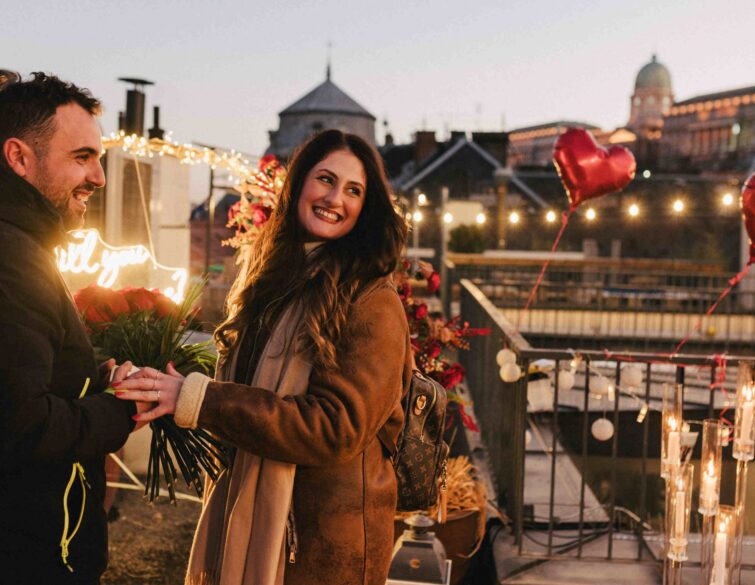 Happy couple during a romantic rooftop proposal with rose petals, candles, and Budapest city view in the background {{brizy_dc_image_alt imageSrc=