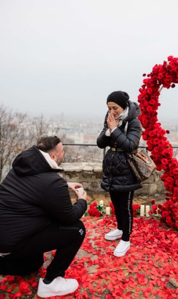 Winter engagement moment at Budapest viewpoint - man proposing under red rose arch with surprised woman in black winter coat and Louis Vuitton bag {{brizy_dc_image_alt imageSrc=