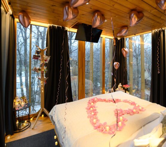 Cozy treehouse interior decorated for proposal with rose petal heart on white bed, rose gold balloons, fairy lights, and winter forest view through panoramic windows {{brizy_dc_image_alt imageSrc=